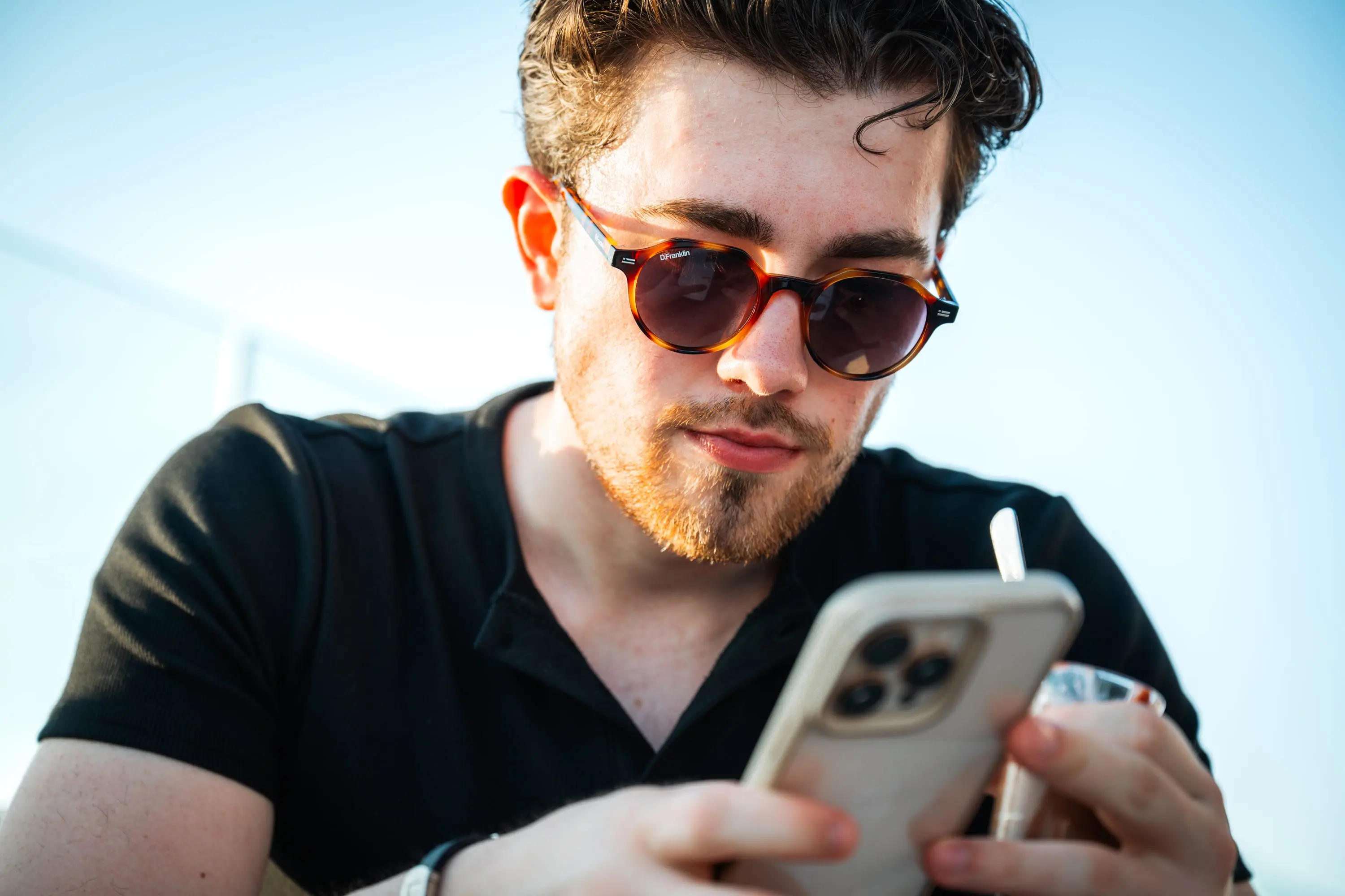 Junger Mann mit Sonnenbrille blickt auf sein Smartphone, Sommerlicht
